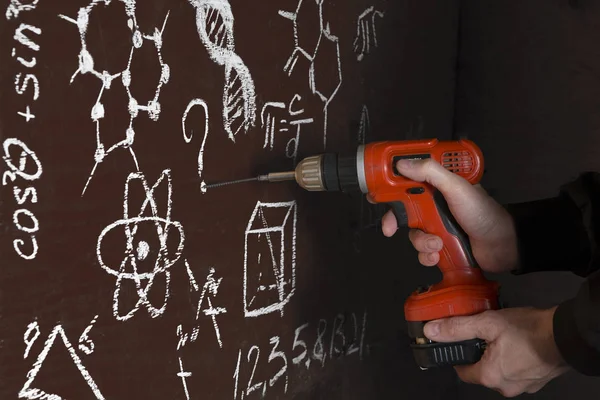 The man from the darkness screws improvised chalkboard using a cordless screwdriver. The board is covered with formulas with a question. Shallow depth of field