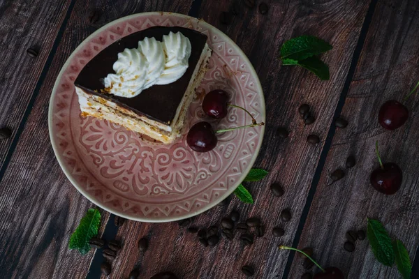 Piece of cake with cream on plate. Sweet cake on wooden background with coffee beans, cherry and mint leaves. Delicious slice of layered tart on plate. Baking and decorating cake