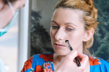 Makyaj yapma süreci. Makyöz yüzüne fırçayla sürmüş. Güzellik salonundaki genç kızıl kadının portresi. Fırçayla makyaj ustasının eli mükemmel cilt ve göz farı yapar.. 