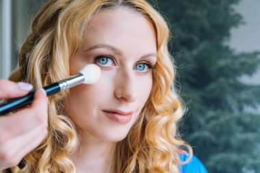 Makyaj yapma süreci. Makyöz yüzüne fırçayla sürmüş. Güzellik salonundaki genç kızıl kadının portresi. Fırçayla makyaj ustasının eli mükemmel cilt ve göz farı yapar.. 