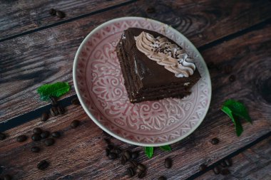 Bir parça kremalı çikolatalı kek. Kahve çekirdekleri ve nane yapraklarıyla ahşap arka planda çikolatalı pasta. Tabakta lezzetli katmanlı tart dilimi. Çikolatalı pasta pişirmek ve dekore etmek