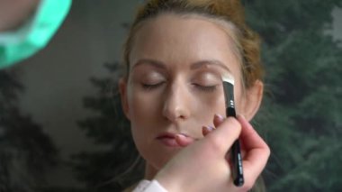 Makyaj yapma süreci. Makyöz yüzüne fırçayla sürmüş. Güzellik salonundaki genç kızıl kadının portresi. Fırçayla makyaj ustasının eli mükemmel cilt ve göz farı yapar. 4k video
