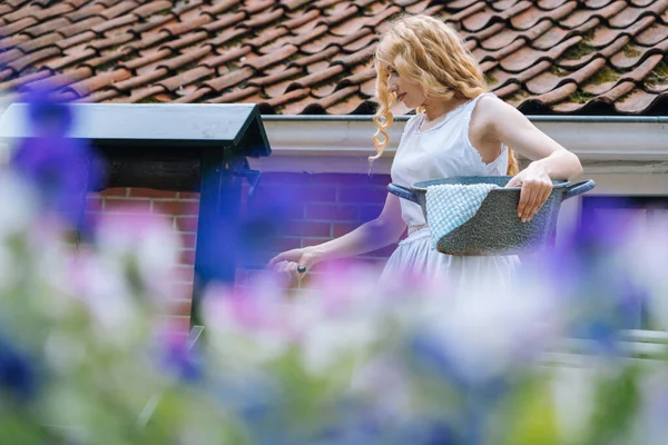 Woman in white dress holding basin near ancient artesian draw-well in village. Colorful violet flowers on foreground. Girl gets water from rural well in summer sunny day for washing. — Stock Photo, Image