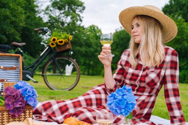 Kareli elbiseli ve şapkalı bir kız örgü örüp piknik battaniyesine oturmuş kitap okuyor ve şarap içiyor. Güneşli bir günde yaz pikniği. Ekmek, meyve, buket çiçekleri. Arka planda bisiklet var. Seçici odak