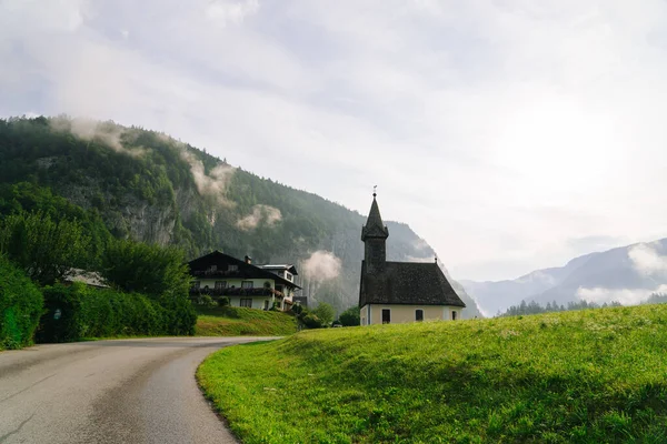 Scenic panoramic view of idyllic church in morning in the alps. Beautiful foggy morning scenery in Alps region, Austria. Great morning view of foggy mountains, fog, house and green meadow in Austria