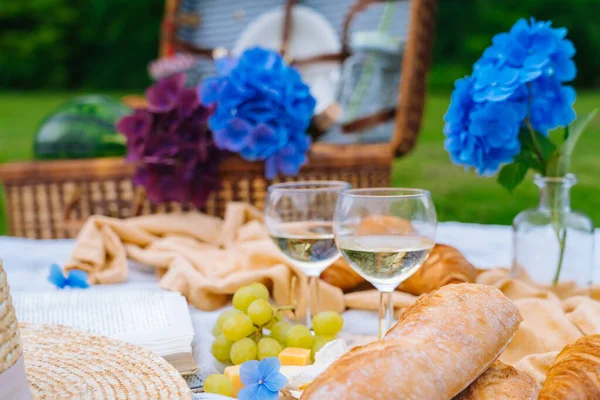 Summer picnic on sunny day with bread, fruit, bouquet hydrangea flowers, glasses wine, straw hat, book and ukulele. Picnic basket on grass with food and drink on white knit blanket. Selective focus.