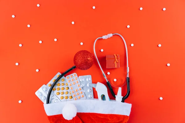 Medicine christmas new year concept banner during coronavirus covid-19 time. Top view of medical pills, disposable protective face mask, gift box, stethoscope in santa hat on red background. CopySpace