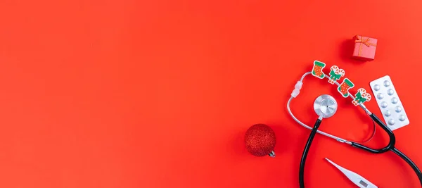 Medicine christmas new year concept banner during coronavirus covid-19 time. Top view of medical pills, disposable protective face mask, gift box, stethoscope in santa hat on red background.