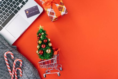 Kredi kartıyla bilgisayardan Noel alışverişi. Yeni yıl dekorasyonları, baston şekerler, alışveriş arabası ve hediye kutusunun üstü kırmızı arka planda yatıyordu. Mutlu Noeller kış tatili satış konsepti.