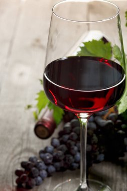 Eski ahşap bir masada bir bardak kırmızı şarap, bir şişe ve kırmızı üzüm..