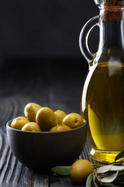 Cam şişede zeytinyağı, seramik kasede taze zeytin ve eski ahşap bir masada zeytin dalı. Koyu arkaplan.