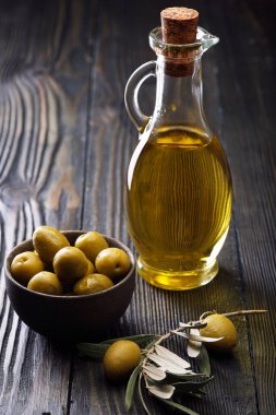 Cam şişede zeytinyağı, seramik kasede taze zeytin ve eski ahşap bir masada zeytin dalı. Koyu arkaplan.