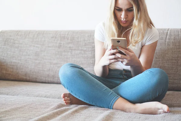 Attraente giovane donna che si rilassa su un divano a casa e utilizza lo smartphone — Foto Stock
