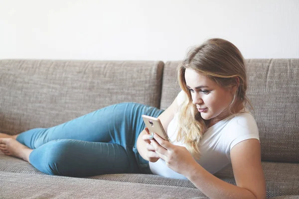 Attraente giovane donna che si rilassa su un divano a casa e utilizza lo smartphone — Foto Stock