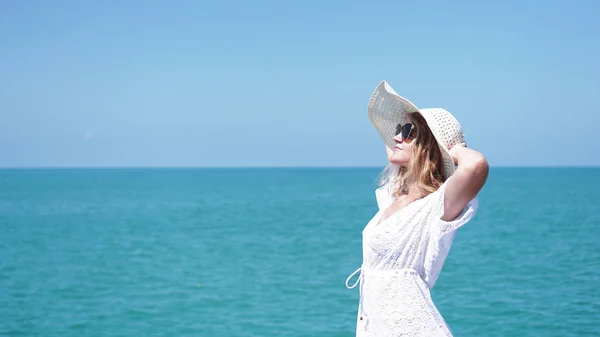 युवा महिला समुद्र के पास रेत पर खड़े और एक टोपी पकड़ते हुए — स्टॉक फ़ोटो, इमेज