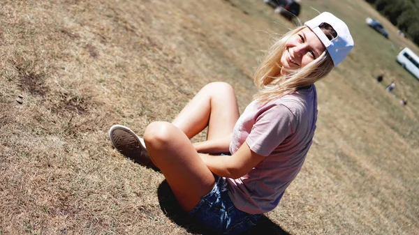 La chica en una gorra y pantalones cortos en las montañas se sienta en la hierba —  Fotos de Stock