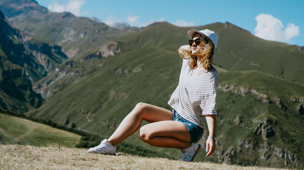 The girl in a cap and shorts in the mountains sits on the grass