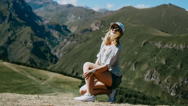 Dziewczyna w czapce i szorty w górach siedzi na trawie — Zdjęcie stockowe