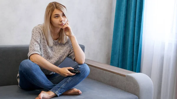 En ung uttråkad kvinna tittar på tv med fjärrkontroll — Stockfoto