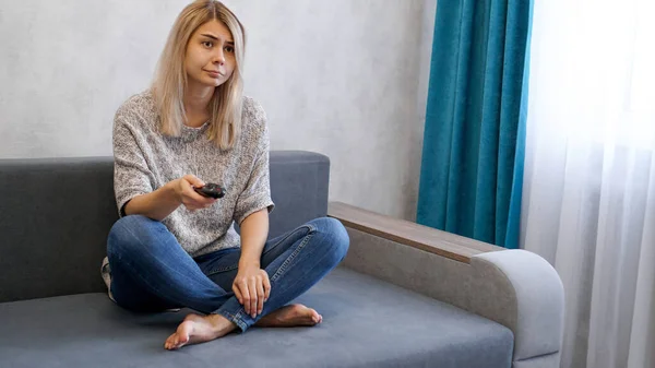 Молодая женщина меняет телеканалы с пультом дистанционного управления, сидя на диване — стоковое фото