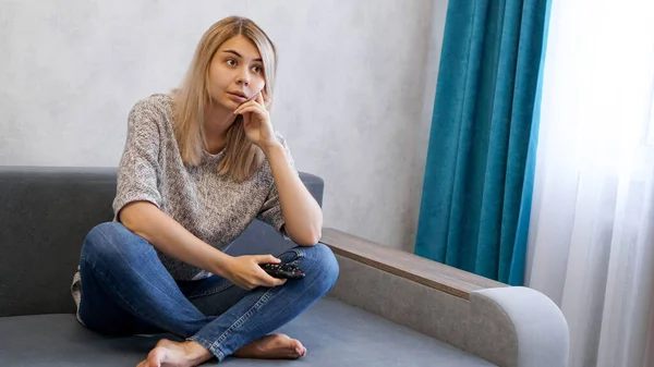 Young woman changing TV channels with remote control sitting on sofa