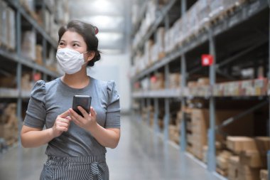 Genç Asyalı kadın market deposunda market arabasıyla günlük kıyafet alışverişi yapıyor. Yeni normal yaşam tarzına sahip. Yüz maskesi takıyor ve Covid19 'dan sonra akıllı telefon kullanıyor.