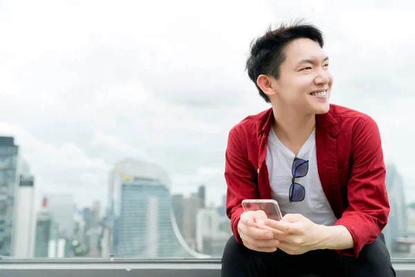 若いアジア人男性カジュアルリラックス赤いシャツ手は都市のダウンタウンのビジネス技術のアイデアの概念を背景にガラスのスマートフォンを保持 — ストック写真