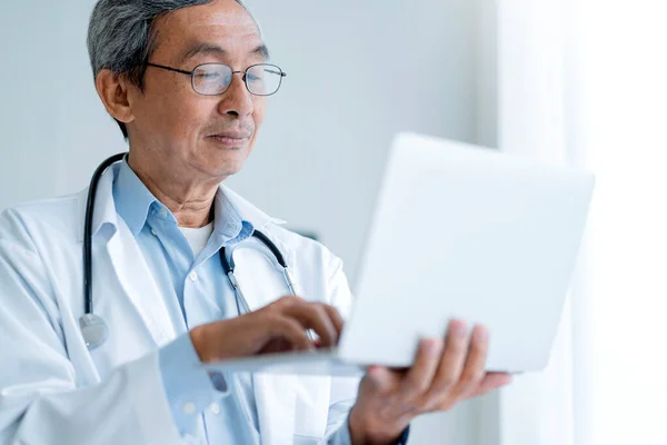 病院でのシニアアジアの男性医師は 医療文書を書き ホワイトクリニックの背景を管理するためのラップトップを使用して — ストック写真