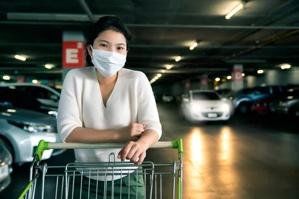 portrait young asian female wear face mask protection hand pull shopping cart alone in parking lot indoor shopping department store mall background