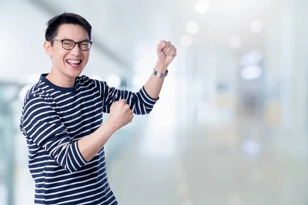 exciting smart casual asian male hand rise big toothy smile with blur office background successful asian male shout and cheerful gesture hand open ready to victory