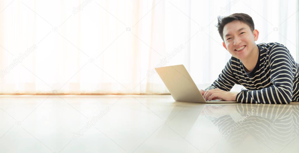 happiness asian smart male smile with confident and joyful working with laptop casual tshirt relax and lay down on wooden floor with home window interior background