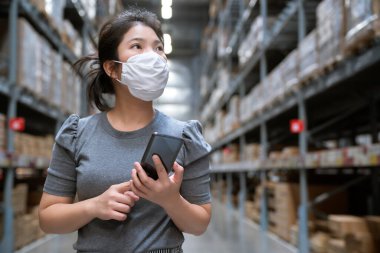 Genç Asyalı kadın market deposunda market arabasıyla günlük kıyafet alışverişi yapıyor. Yeni normal yaşam tarzına sahip. Yüz maskesi takıyor ve Covid19 'dan sonra akıllı telefon kullanıyor.