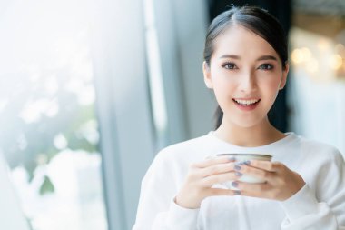 Girişimci girişimi sahibi çekici Asyalı kadın iş kadını akıllı telefon ve dizüstü bilgisayarla iletişim kuruyor beyaz elbiseler giyip güvenle gülümsüyor ve ofis geçmişi bulanıklaşıyor.