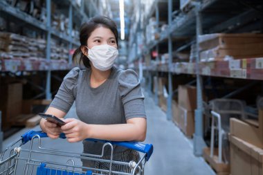 Genç Asyalı kadın market deposunda market arabasıyla günlük kıyafet alışverişi yapıyor. Yeni normal yaşam tarzına sahip. Yüz maskesi takıyor ve Covid19 'dan sonra akıllı telefon kullanıyor.
