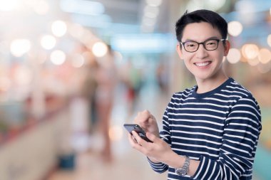akıllı tesadüfi Asyalı mutluluk erkek gözlük takar gülümseme ve neşeli el telefonu kullanır alışveriş merkezi soyut bulanık arka plan teknoloji iletişim fikirleri içeren satın alma düğmesine basmaya hazır.