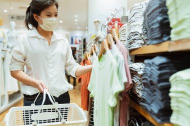 Covid salgından sonra yeni normal. Asyalı genç bayan, mağaza arka planında yeni bir yaşam tarzı olan butik dükkanında maske ya da giysi giyiyor.
