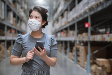 Genç Asyalı kadın market deposunda market arabasıyla günlük kıyafet alışverişi yapıyor. Yeni normal yaşam tarzına sahip. Yüz maskesi takıyor ve Covid19 'dan sonra akıllı telefon kullanıyor.