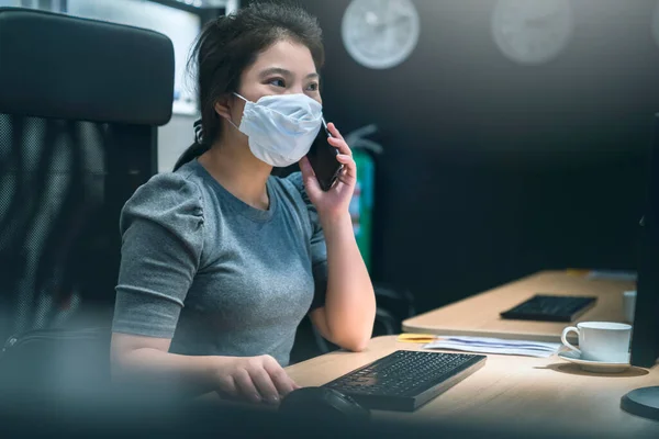 young asian female wear facemask protection while working hard busy multi task with smartphone and table computer late night new normal work lifestyle in office