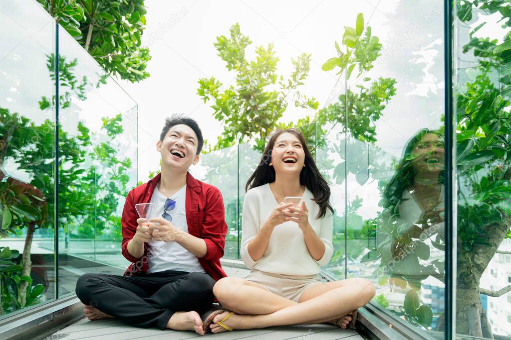 attractive asian couple casual dress enjoy cheerful laugh and smile hand play online mobile game together competition on smartphone together in modern garden