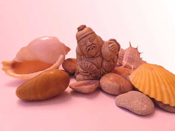 Японская Фигурка Старик Рыбой Руках Среди Раковин Камней Розовом Фоне — стоковое фото