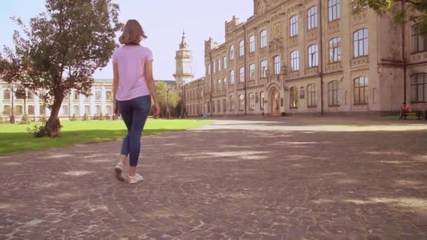 背面の金髪の学生キャンパス エリアに歩いて ピンクの シャツとジーンズを散策 背景を身に着けている金髪の女の子美しい古い大学 — ストック動画