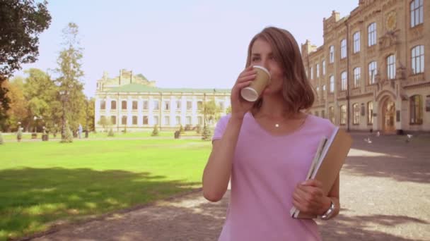 Giovane studentessa bionda che beve caffè passeggiando dopo la lezione vicino all'università. ragazza con i capelli biondi indossa rosa t-shirt e jeans passeggiata sullo sfondo bellissimo vecchio collegio — Video Stock