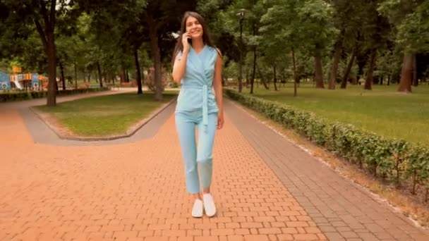 Jovem caucasiano mulher falando com amigo por móvel andando na rua no parque da cidade — Vídeo de Stock