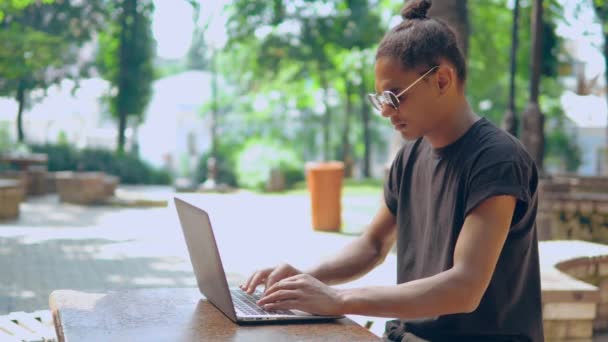 Профіль молодий хіпстер в сонцезахисних окулярах, використовуючи ноутбук на відкритому повітрі — стокове відео