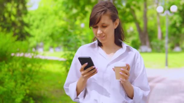 Young businesswoman chatting online drinking coffee stroll in park — Stock Video