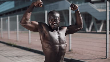Poz çıplak gövde ile atletik Afrikalı-Amerikalı adam açık havada kasları gösterir. Siyah adam stadium yanında poz kasları gösterir. Vücut geliştirme, poz, sağlık, fitness, güzellik.