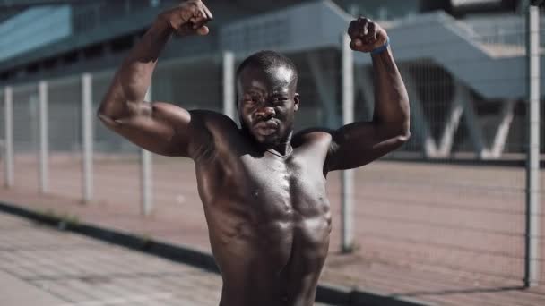 Homem afro-americano atlético com um tronco nu posando mostra seus músculos ao ar livre. Homem negro posando perto do estádio, mostra seus músculos. Musculação, posando, saúde, fitness, beleza . — Vídeo de Stock