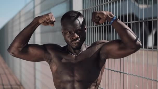 Athletic African American człowiek nagi tors pozowanie pokazuje jego mięśnie na zewnątrz. Czarny człowiek pozowanie w pobliżu stadionu, pokazuje jego mięśnie. Kulturystyka, pozowanie, zdrowie, fitness, uroda. — Wideo stockowe