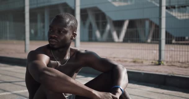 Retrato del atlético afroamericano que mira a la cámara después del entrenamiento.El joven musculoso se sienta en un tapete cerca del estadio deportivo. Fitness, salud, deportes, musculación, estilo de vida activo — Vídeo de stock