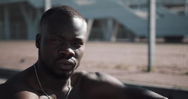 Ritratto di uomo afroamericano atletico che guarda nella macchina fotografica dopo l'allenamento.Giovane uomo muscoloso si siede su un caremat vicino allo stadio sportivo. Fitness, salute, sport, bodybuilding, stile di vita attivo — Video Stock